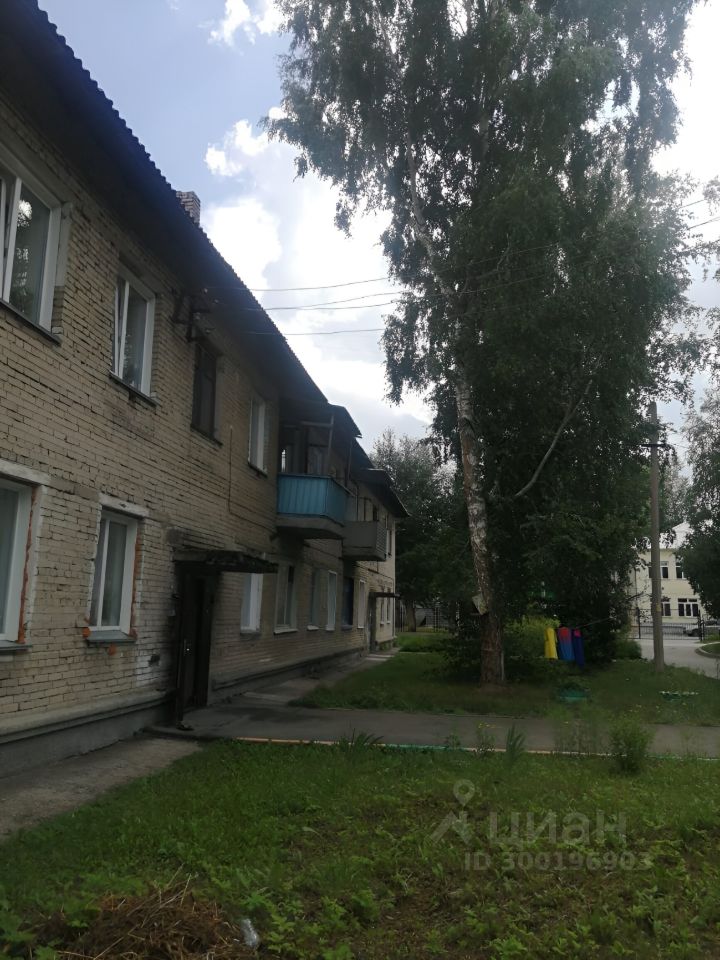 Здание выполнено в советском архитектурном стиле с кирпичной отделкой. На фасаде видны окна с простыми рамами и балкон, обшитый металлическим каркасом и окрашенный в голубой цвет. У входа расположена простая дверь под навесом. Вокруг здания ухоженная территория с газоном и небольшими кустами. Цвет стен бежевый, с частичными следами старой краски. Полы и дорожки выполнены из бетона, что создает практичное и функциональное пространство. Общий вид здания создает ощущение простоты и практичности, характерное для жилых комплексов того времени.
