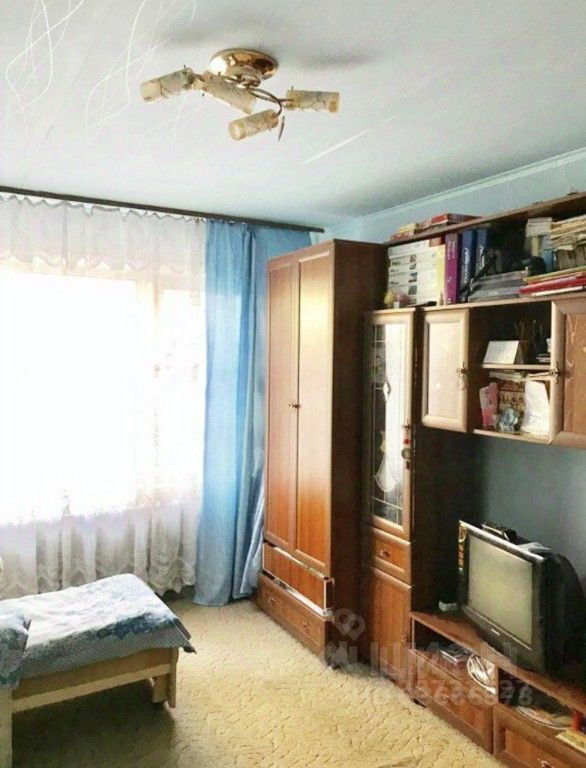 Комната оформлена в простом и функциональном стиле. На стенах светло-голубой цвет, потолок ровный с круглой люстрой. Окно обрамлено легкими шторами, создающими уютное освещение. В углу стоит деревянный шкаф с распашными дверцами, а рядом размещен телевизор на низком тумбочке. По центру комнаты расположена кровать с простыней в голубых тонах. Пол покрыт мягким ковролином, что добавляет тепла и уюта. Общая атмосфера комнаты спокойная и комфортная, идеально подходящая для отдыха.