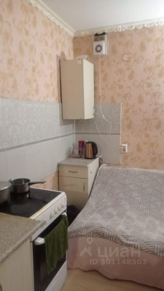 Кухня оформлена в светлых тонах с цветочным узором на стенах. На видимой части кухни расположены газовая плита с варочной панелью, кастрюля и полотенце. Рядом стоит столешница с электрочайником и несколькими кухонными принадлежностями. В углу находится шкаф для хранения, выполненный в светлом цвете. Полы покрыты светлым ламинатом. Общий стиль интерьера создает уютную атмосферу, подчеркивая функциональность пространства.