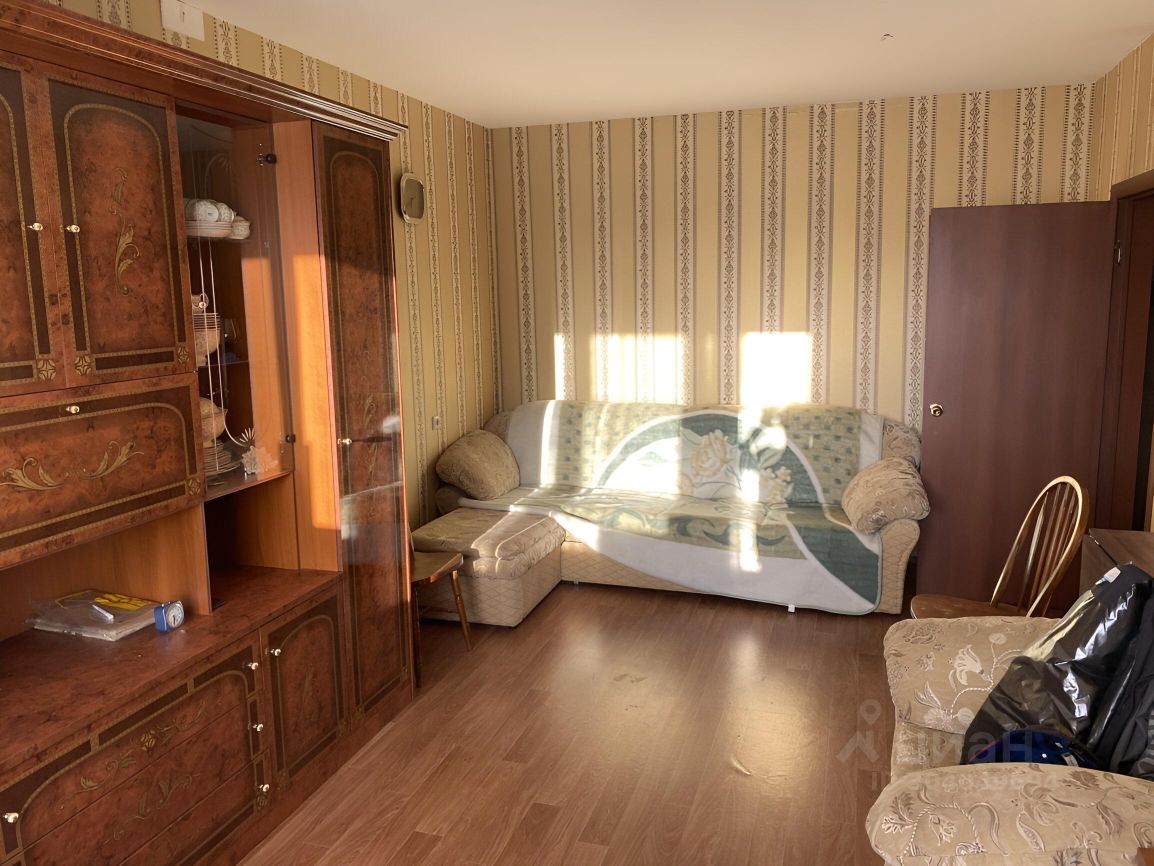 Комната оформлена в классическом стиле с элементами деревянной мебели. На стенах присутствует обои с вертикальным рисунком, создающие ощущение высоты. В центре находится диван с мягкими подушками и покрывалом, а также кресло с узорчатой обивкой. Напольное покрытие выполнено из ламината, что придаёт уют. Стенка с витриной и шкафом из темного дерева добавляет элегантности. Освещение проникает через окно, создавая теплую атмосферу. В комнате также виден столик, что делает пространство функциональным для отдыха и общения.