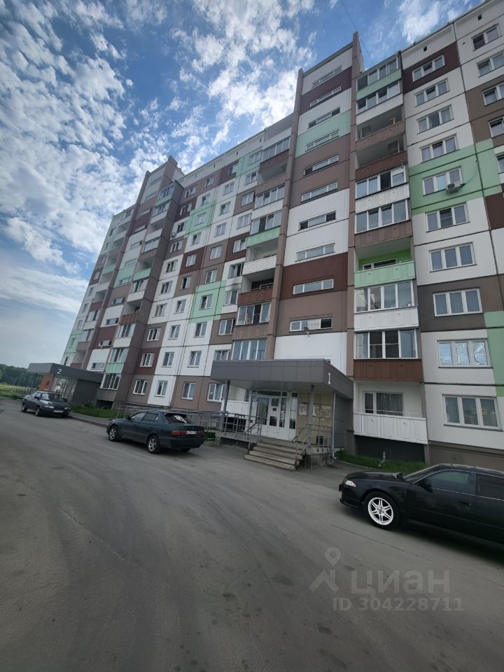 Здание выполнено в современном архитектурном стиле с элементами панельного домостроения. Фасад имеет разнообразные цвета: коричневый, зеленый и белый, что придает ему привлекательный вид. На переднем плане видна входная группа с лестницей и перилами, а также окна с балконами, что свидетельствует о функциональном дизайне. Структурные особенности включают большие окна, обеспечивающие естественное освещение, и четкие линии, создающие ощущение современности. Материалы отделки простые, с гладкой поверхностью, что подчеркивает практичность и доступность жилья.