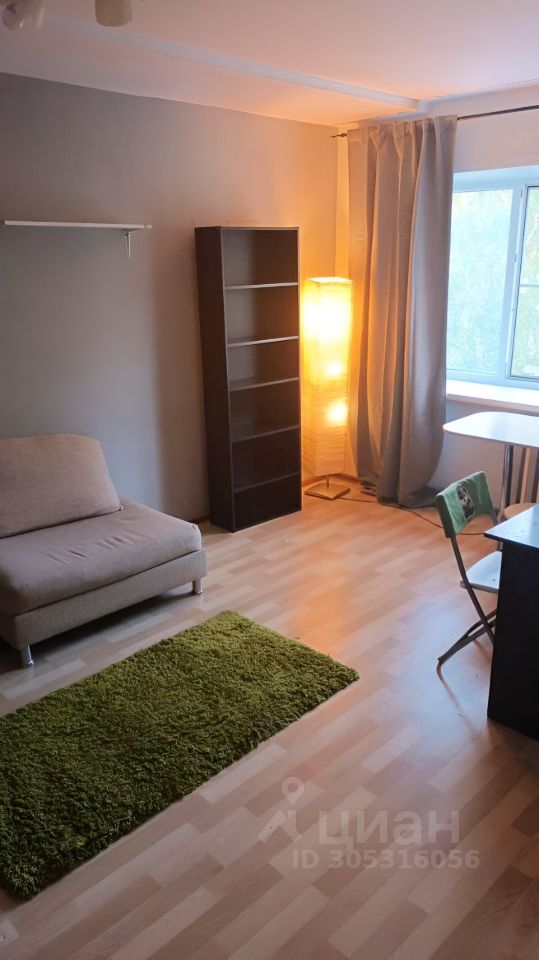 Комната оформлена в современном стиле с нейтральными цветами. На полу светлый ламинат, а в центре расположен зеленый коврик. В углу стоит мягкий диван бежевого цвета, рядом с ним высокая книжная полка черного цвета. У окна установлена светлая занавеска, а рядом с ней стоит стильный торшер, создающий уютное освещение. В комнате также имеется рабочий стол с металлическими ножками и складной стул. Пространство выглядит светлым и просторным, идеально подходящим для отдыха или работы.