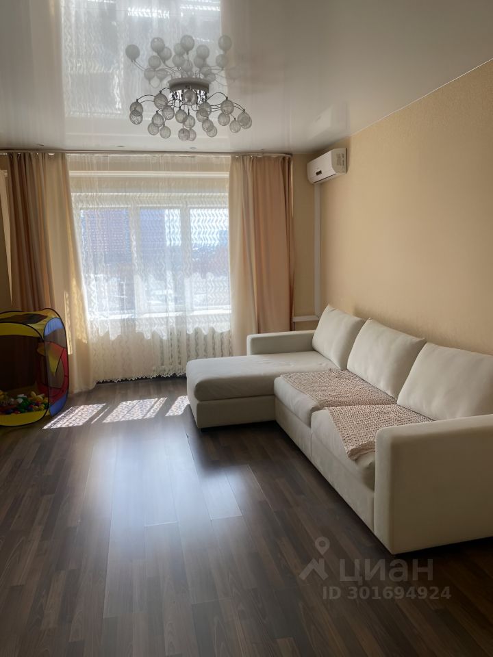 Комната оформлена в современном стиле с элементами минимализма. На стенах светлые тона, а пол выполнен из темного ламината, создающего контраст. В центре потолка расположена стильная люстра с округлыми стеклянными элементами. Большие окна, обрамленные легкими шторами, обеспечивают естественное освещение и создают уютную атмосферу. Угловой диван светлого цвета с текстильным покрывалом дополняет пространство, а рядом находится детский игровой уголок с ярким игровым домиком. Кондиционер установлен на стене, что добавляет комфорт в теплое время года.