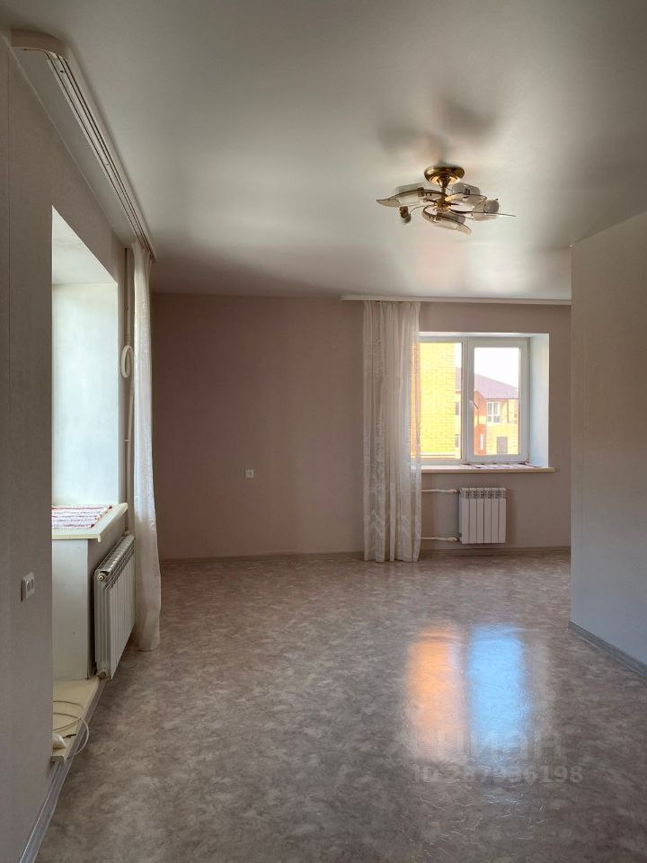 Помещение оформлено в современном стиле с нейтральными цветами стен, выполненными в светло-песочном оттенке. Пол имеет гладкую, блестящую поверхность, создающую ощущение простора. Окна большие, с легкими занавесками, обеспечивающими естественное освещение. В углу расположено радиаторное отопление, что добавляет уют. Потолок ровный, с потолочным светильником, который гармонично дополняет интерьер. Комната выглядит просторной и светлой, что делает её универсальным пространством для различных целей.