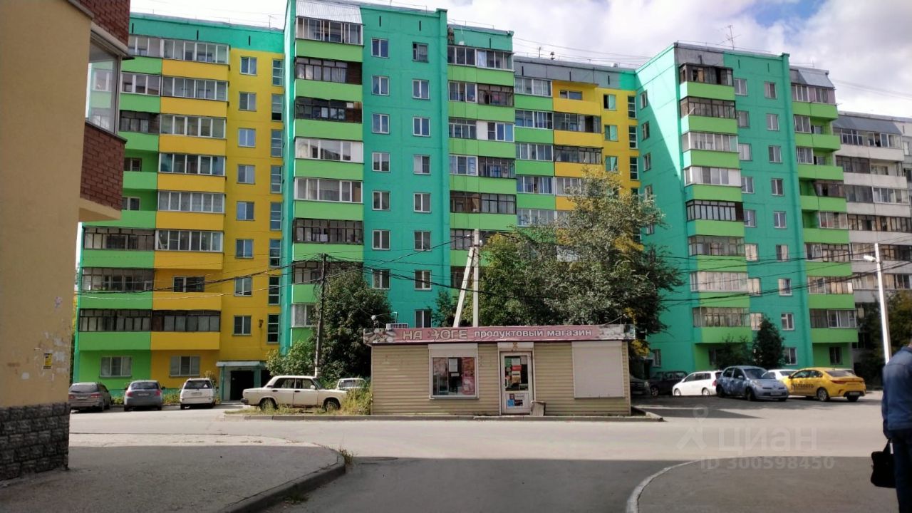Здание выполнено в современном архитектурном стиле с яркой цветовой гаммой, сочетающей зеленые, желтые и голубые оттенки. Фасад имеет множество окон, что обеспечивает хорошее естественное освещение. Структурные особенности включают балконы и большие витражные окна. Визуально заметны элементы, характерные для многоквартирных домов, такие как горизонтальные линии и симметричное расположение окон. Используемые материалы выглядят современно, с акцентом на практичность и долговечность. Цвета фасада создают жизнерадостное и привлекательное впечатление, что делает здание заметным в городской среде.