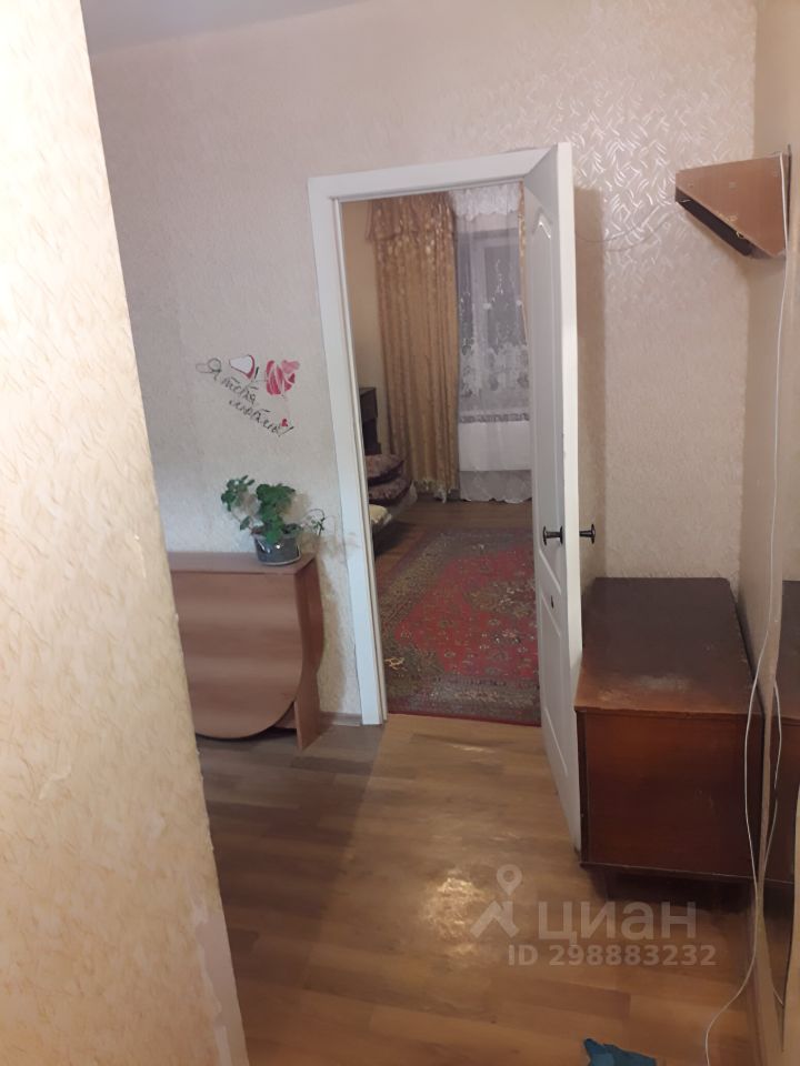Видна прихожая с простым дизайном, стены окрашены в светлый цвет с текстурой. Пол выполнен из светлого дерева, создавая уютную атмосферу. Слева расположена деревянная тумба, а справа — комод. В открытой двери виден соседний зал с ковром и длинными шторами, что добавляет тепла и уюта. Освещение мягкое, создающее комфортное пространство. Растение в горшке на тумбе придаёт живость интерьеру, а общая отделка помещения выглядит аккуратно и стильно.