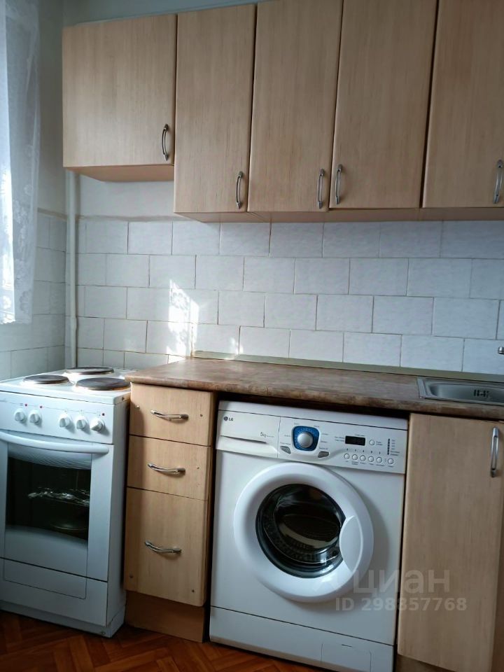 Кухня с современным дизайном, выполненная в светлых тонах. Верхние шкафчики из светлого дерева обеспечивают достаточное пространство для хранения. На столешнице из дерева расположены плита и стиральная машина, что указывает на функциональность пространства. Стены облицованы белой плиткой, создающей чистый и аккуратный вид. Полы выполнены из паркета, добавляя теплоту в интерьер. В комнате присутствует естественное освещение благодаря окну с занавесками, что делает пространство более уютным и приветливым.