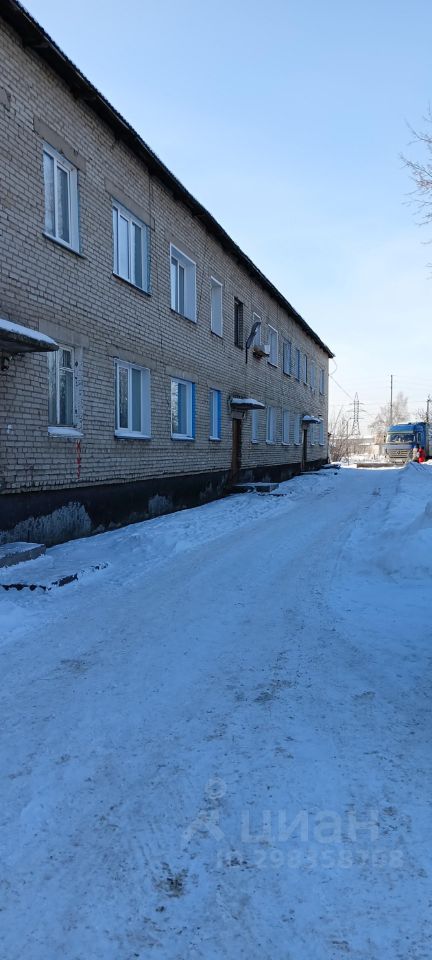Здание выполнено в простом архитектурном стиле с кирпичной отделкой, что придаёт ему классический вид. Фасад имеет ряд окон, оформленных белыми рамами, что создает контраст с серым цветом кирпичей. Структурные особенности включают двухэтажное строение с плоской крышей и небольшими выступами. На переднем плане видна заснеженная дорожка, ведущая к входу. Основные материалы — кирпич и дерево, что обеспечивает прочность и долговечность. Цветовая гамма включает серые и белые оттенки, создавая спокойную и нейтральную атмосферу.
