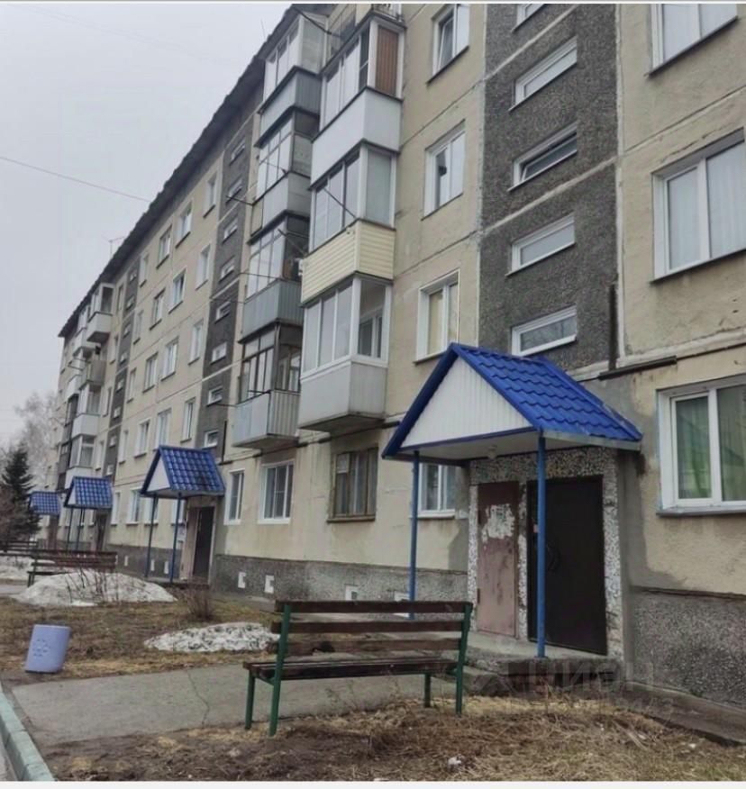 Здание выполнено в советском архитектурном стиле с характерными элементами, такими как балконы и простые формы. Фасад окрашен в светлые тона, с серыми вставками и большими окнами. Входная зона оформлена крышей с голубой черепицей и поддерживается столбами. Видны лавочки для отдыха, расположенные вдоль тротуара, что создает уютную атмосферу. Используемые материалы включают бетон и металл, а цветовая гамма включает серые, белые и голубые оттенки, что придает зданию современный вид, сохраняя при этом элементы традиционного дизайна.