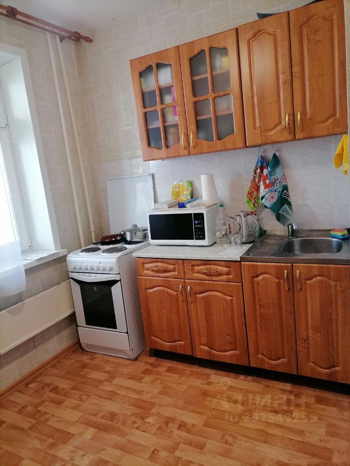 Кухня выполнена в простом и функциональном стиле. На стенах светлая плитка, а на полу — деревянное покрытие. Мебель представлена деревянными шкафчиками с витражными дверцами и столешницей из светлого материала. В углу расположена газовая плита и микроволновая печь. Рядом находится раковина с металлической поверхностью. Окно с белыми шторами обеспечивает естественное освещение, создавая уютную атмосферу. Яркие кухонные полотенца добавляют цвет в обстановку, а открытые полки содержат посуду и кухонные принадлежности, подчеркивая практичность пространства.