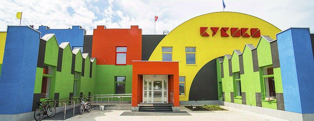 Архитектурный стиль здания характеризуется яркой, современно оформленной фасадной частью с элементами геометрического дизайна. Структурные особенности включают ассиметричные формы и различные уровни, создающие динамичный вид. Основная зона представлена входной группой с широкими стеклянными дверями и яркими цветными стенами, выполненными в оттенках красного, зеленого и синего. Используемые материалы включают гладкую штукатурку и стекло, что придает зданию современный и привлекательный вид. Цветовая палитра создает жизнерадостную атмосферу, акцентируя внимание на оригинальности архитектурного решения.