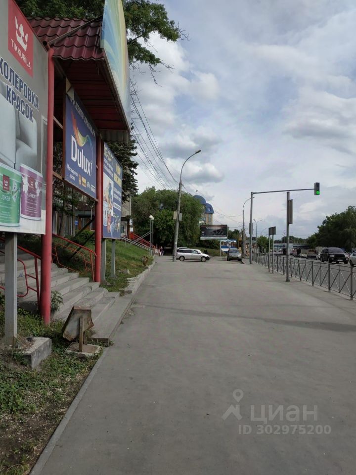 На фотографии виден тротуар с асфальтовым покрытием, который ведет к зданию с красной крышей и рекламными щитами. Ступени из бетона ведут к входу, обрамленному металлическими перилами. Рядом расположены рекламные баннеры с изображениями красок и строительных материалов. На заднем плане видны деревья и автомобили, а также светофор. Цветовая гамма включает серые и красные оттенки, создавая контраст с зелеными насаждениями. Пространство выглядит организованным и доступным для пешеходов.
