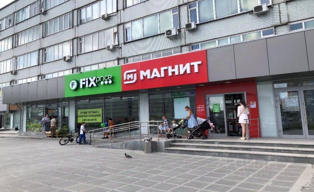 Фасад здания выполнен в современном стиле с элементами минимализма. На переднем плане расположены входы в магазины с яркими вывесками: зелёная вывеска FIX price и красная вывеска Магнит. Структурные особенности включают стеклянные витрины, что позволяет видеть внутреннее пространство. Входы оформлены ступенями и пандусом, что обеспечивает доступность. Основные материалы – стекло и металл, цвета фасада нейтральные, с акцентом на яркие элементы вывесок. Общая атмосфера пространства выглядит дружелюбной и доступной для посетителей.