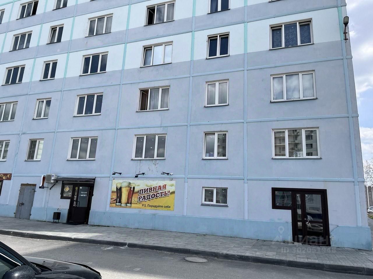 Фасад здания выполнен в современном стиле с элементами минимализма. Структура включает несколько окон с прямыми линиями, расположенных симметрично. Основной цвет — светло-голубой, дополненный белыми рамами окон. Входная группа оформлена в темных тонах, что создает контраст с общим цветом здания. На стене вывеска с изображением пива, что указывает на коммерческое назначение нижнего этажа. Материалы отделки фасада выглядят современно, с гладкой текстурой. Отсутствие балконов придает зданию строгий и упорядоченный вид.