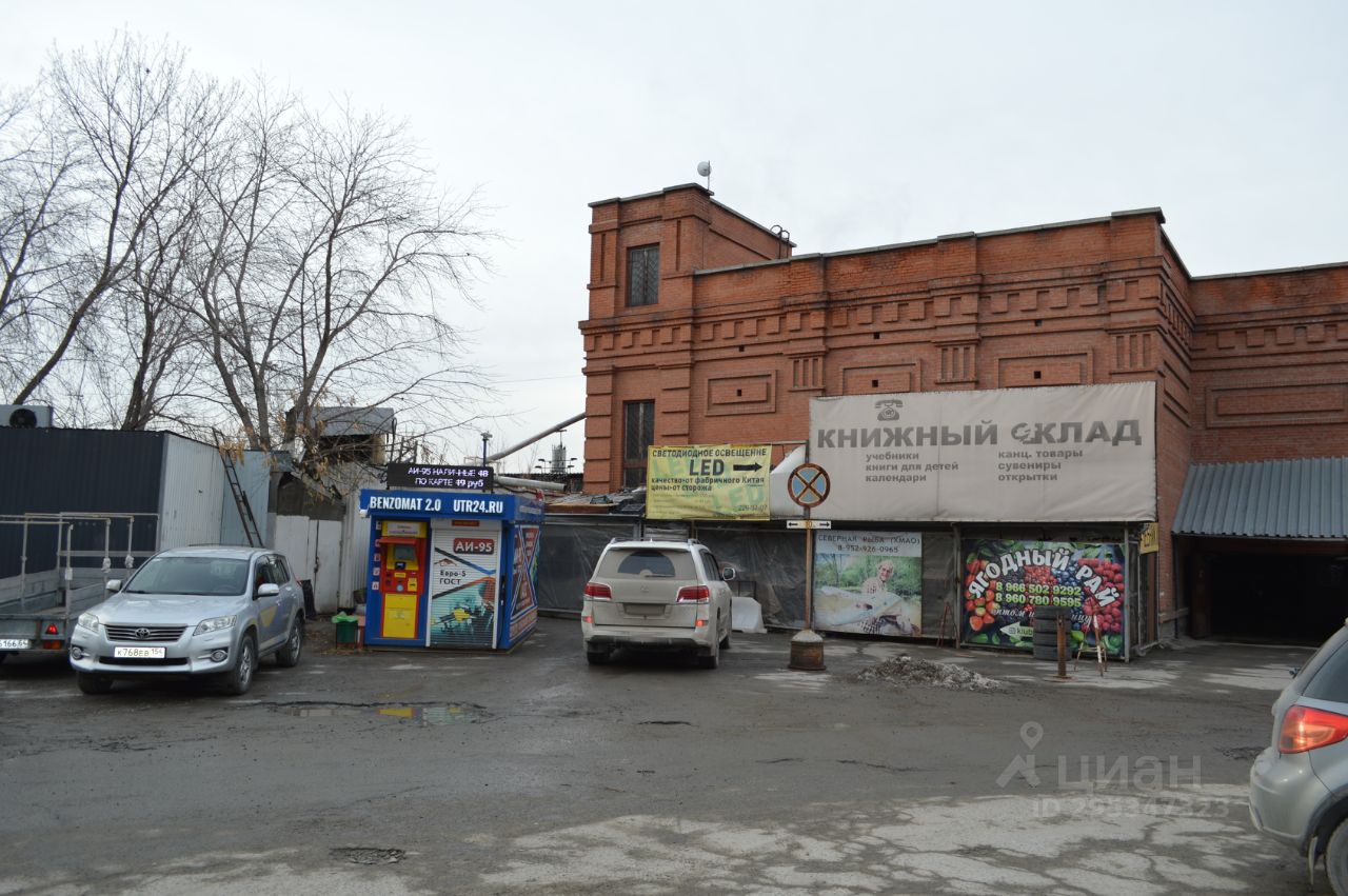 Здание выполнено в кирпичном стиле с характерными архитектурными элементами, такими как рельефные украшения и большие окна. На фасаде видны вывески с названиями магазинов, включая 'Книжный склад'. Структурные особенности включают несколько уровней и крытую часть, которая может служить входом. Основные цвета – кирпичный красный и серый, с элементами рекламы, добавляющими яркие акценты. Материалы, использованные в отделке, создают индустриальный вид, подчеркивая функциональность пространства.