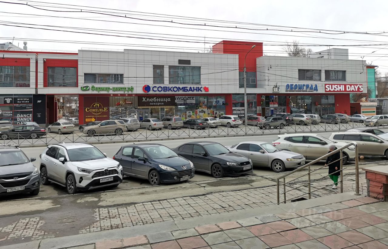 Фасад здания выполнен в современном архитектурном стиле с элементами минимализма. Видны большие витрины, которые обеспечивают естественное освещение и привлекают внимание к коммерческим помещениям. Структурные особенности включают несколько этажей с различными магазинами и ресторанами на первом этаже. Основные цвета фасада — белый, красный и серый, что создает контраст и динамичность. Материалы отделки — стекло, металл и бетон, придающие современный вид. Пространство организовано для удобного доступа, с пешеходными дорожками и парковкой для автомобилей.