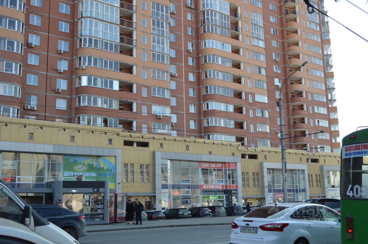 Здание выполнено в современном архитектурном стиле с элементами фасадного остекления. Видны многоквартирные этажи с балконами, обрамленными металлическими перилами. На первом этаже расположены коммерческие помещения с большими витринами, что создает открытое пространство. Цветовая гамма включает теплые оттенки кирпича и светлые тона, подчеркивающие современный дизайн. Используются стеклянные и металлические материалы, что придает зданию стильный и функциональный вид. Общая композиция создает ощущение динамики и современности жилого комплекса.