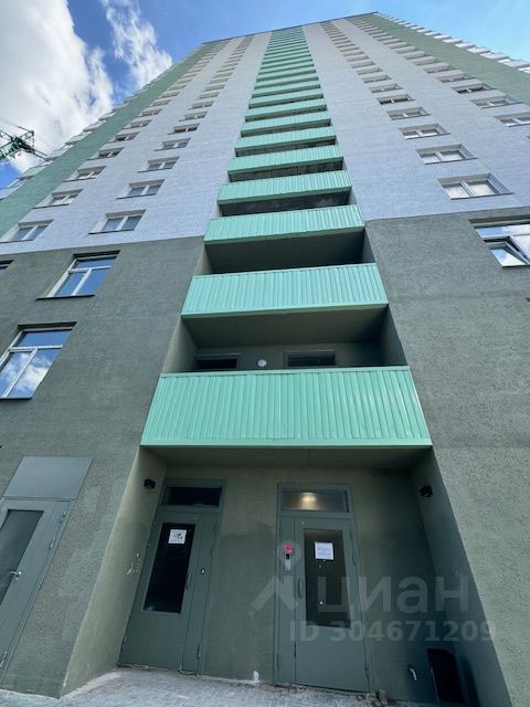 Фасад здания выполнен в современном архитектурном стиле с элементами минимализма. Структурные особенности включают вертикальные линии и симметричное расположение окон, что создает ощущение высоты. На первом этаже расположены входные двери, окрашенные в серый цвет, и балконы с зелеными панелями, придающими яркость. Цветовая палитра включает светло-серый и зеленый оттенки, а отделка выполнена из современных материалов, что подчеркивает стиль и функциональность. Окна имеют прямоугольную форму, обеспечивая достаточное количество естественного света.