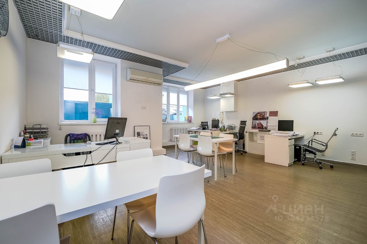 Офисное пространство с современным дизайном, выполненное в светлых тонах. Основное внимание привлекает длинный стол с белой столешницей и пластиковыми стульями. Вдоль стен расположены окна, пропускающие естественный свет. На стенах могут быть размещены картины и документы. Пол выполнен из деревянных панелей, что создает уютную атмосферу. В помещении также видны офисные принадлежности и техника, включая компьютер и принтер. Освещение обеспечивается современными светильниками, создающими комфортные условия для работы.