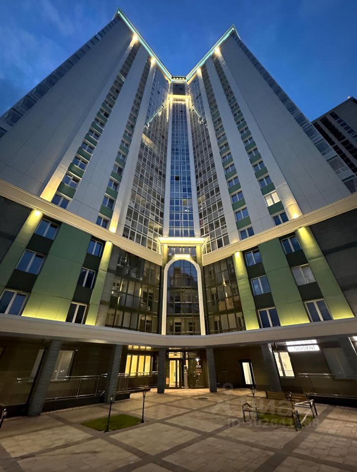 Здание выполнено в современном архитектурном стиле с элементами минимализма. Главной особенностью является высокая стеклянная фасадная часть, которая отражает свет и создает эффект открытости. Внутреннее пространство оформлено в светлых тонах, с использованием плитки и современных отделочных материалов. На первом этаже расположены входные группы с большими окнами, обеспечивающими естественное освещение. Арочные элементы и аккуратные линии добавляют элегантности. Площадка перед входом вымощена плиткой, создавая уютное пространство для отдыха.