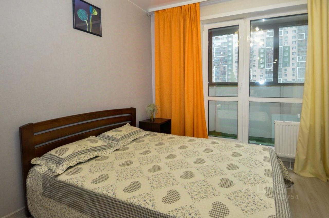 Спальня оформлена в современном стиле с элементами минимализма. Основным акцентом является двуспальная кровать с деревянным каркасом и текстильным покрывалом с узором в виде сердечек. Окно, обрамлённое яркими оранжевыми шторами, обеспечивает естественное освещение. Рядом с кроватью расположен небольшой прикроватный столик с вазой, что добавляет уюта. Стены окрашены в светлый оттенок, что визуально расширяет пространство. Пол выполнен из светлого материала, создающего гармоничное сочетание с общим дизайном комнаты.