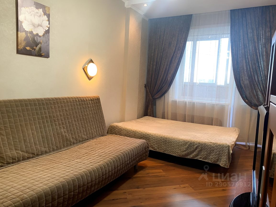 Комната оформлена в современном стиле с мягкими цветами и уютной атмосферой. На стенах светлые обои, а на одной из них висит картина с цветком. Виден угловой диван с текстурированной обивкой, который может служить как место для отдыха. Рядом расположена кровать с простыней, создающая ощущение комфорта. Окно, обрамленное тяжелыми шторами, пропускает мягкий свет, добавляя тепла в интерьер. Пол покрыт паркетом, что придает комнате элегантный вид. Освещение обеспечивается современным настенным светильником, подчеркивающим детали дизайна.