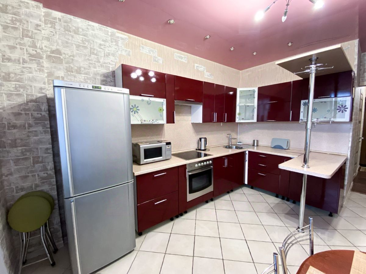 Кухня оформлена в современном стиле с яркими красными фасадами шкафов и светлыми столешницами. Стены отделаны плиткой под кирпич и обоями, создающими уютную атмосферу. В угловой части расположена современная техника, включая холодильник и микроволновую печь. Пол выложен плиткой, что облегчает уборку. Обеденная зона представлена небольшим столом и стульями, что делает пространство функциональным. Освещение обеспечивается встроенными светильниками, создающими мягкий свет. Общая палитра включает красные, серые и светлые оттенки, что придает кухне стильный и современный вид.