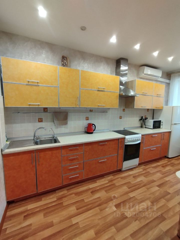 Кухня оформлена в современном стиле с яркими элементами. Верхние шкафчики выполнены в желтом цвете с металлическими ручками, а нижние имеют оранжевый оттенок. Рабочая поверхность из светлого материала с мойкой и кухонной плитой. В углу расположена духовка, а рядом — микроволновая печь. Пол покрыт ламинатом с теплым древесным оттенком. Освещение обеспечивается встроенными светильниками, создающими уютную атмосферу. Стены оформлены нейтральным цветом с текстурой, что придает пространству современный вид. Кухонные аксессуары, такие как чайник, добавляют функциональности.