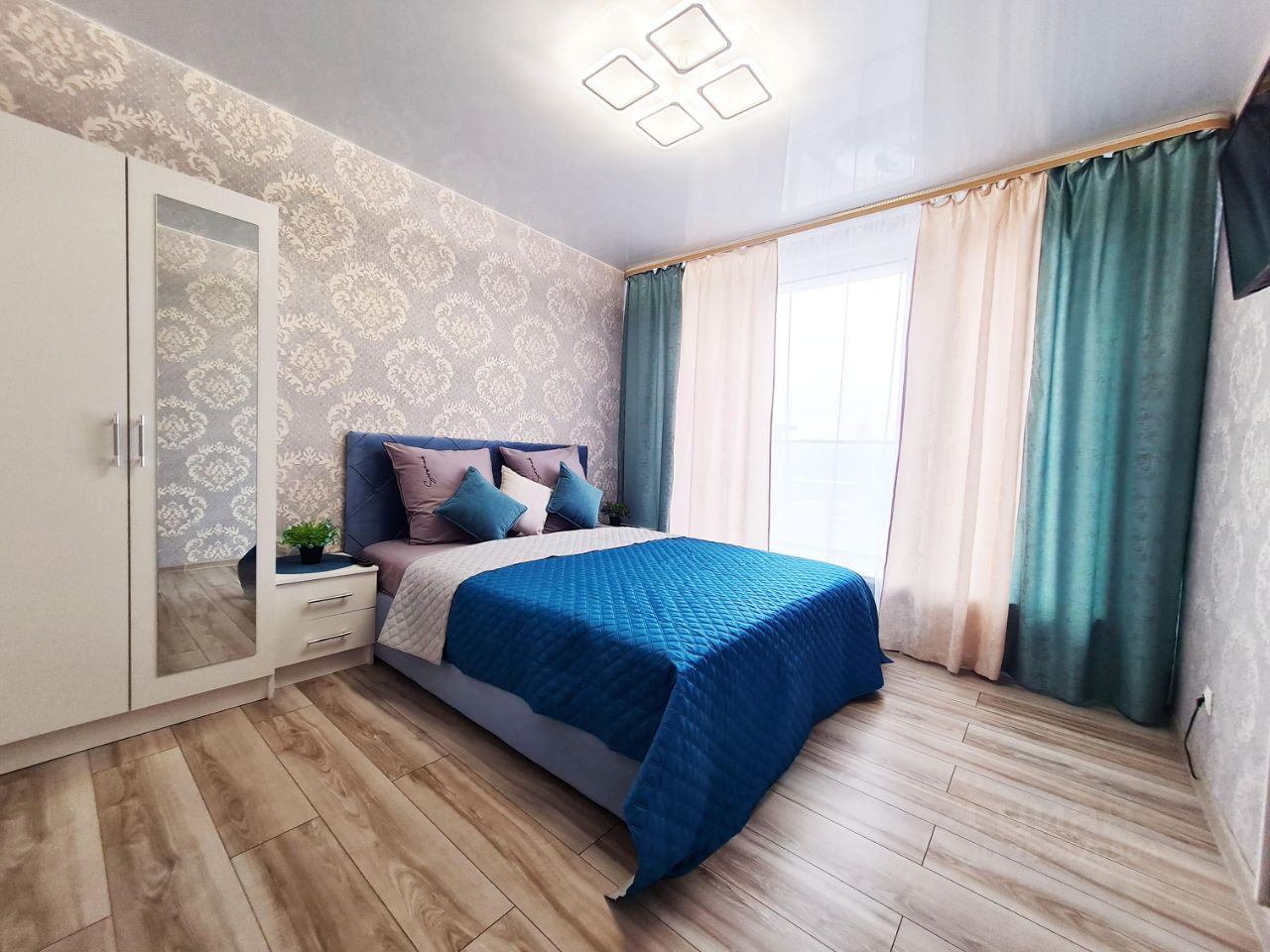 Современная спальня с элегантным дизайном. Стены украшены светлым обоями с изысканным узором. Кровать с голубым покрывалом и мягкими подушками создает уютную атмосферу. Рядом находится белый комод с зеркалом и небольшой горшок с растением. Окна обрамлены шторами в нежных пастельных тонах, добавляющими мягкость в интерьер. Пол выполнен из светлого дерева, что визуально расширяет пространство. Освещение представлено стильной потолочной люстрой, создающей комфортный свет. В комнате царит гармония и спокойствие, идеально подходя для отдыха.