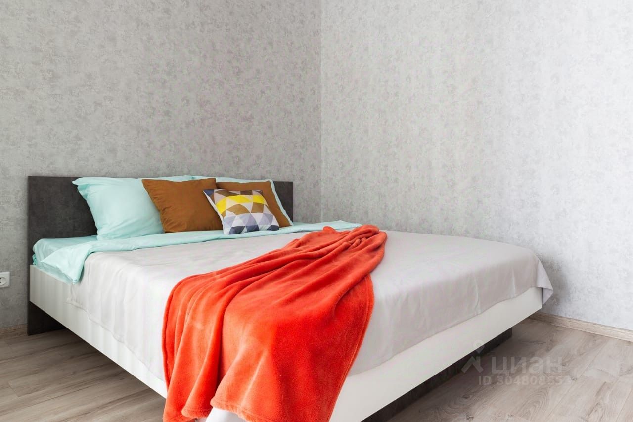 В комнате представлена современная спальня с минималистичным дизайном. Кровать с белым основанием и темной спинкой украшена светлыми подушками и одеялом яркого оранжевого цвета. Стены отделаны светло-серой текстурированной обоями, создающими уютную атмосферу. Пол выполнен из светлого дерева, что добавляет тепла в интерьер. На кровати расположены декоративные подушки с геометрическим узором, которые придают пространству стильный акцент. Общее оформление комнаты создает ощущение простоты и комфорта.