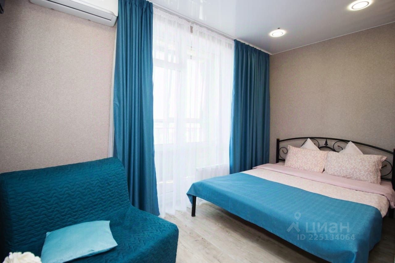 Комната оформлена в современном стиле с акцентом на уют и комфорт. На стенах светлое покрытие, а пол выложен светлым деревом. В центре находится двуспальная кровать с мягким текстилем, украшенная подушками с цветочным принтом. Рядом расположен диван глубокого бирюзового цвета, создающий гармонию с занавесками того же оттенка. Окно обрамлено легкими прозрачными шторами, позволяющими естественному свету проникать в комнату. Освещение представлено встроенными светильниками, создающими мягкую атмосферу. Пространство выглядит аккуратно и стильно, идеально подходя для отдыха.