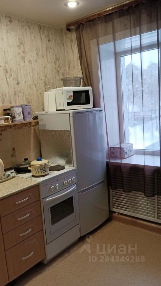 Кухня выполнена в современном стиле с элементами минимализма. Основные характеристики включают встроенные шкафчики из светлого дерева и рабочую поверхность из светлого камня. На кухне расположены газовая плита с духовкой, микроволновая печь и холодильник, выполненные в серебристом цвете. Окно с прозрачными занавесками пропускает свет, создавая уютную атмосферу. Пол покрыт светлым линолеумом, а стены оформлены обоями с тонким узором. В углу находится радиатор, обеспечивающий комфортную температуру.