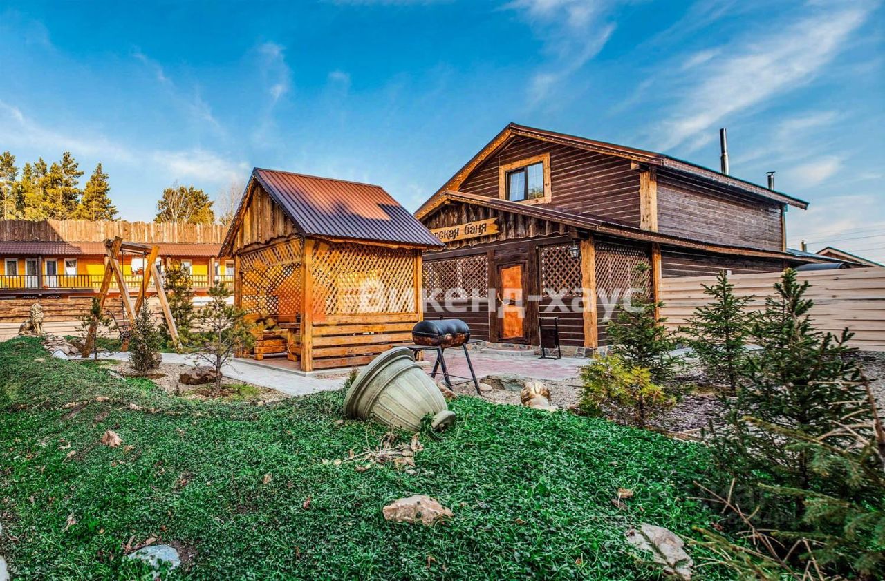 Деревянный дом в традиционном русском стиле с элементами rustic. На переднем плане расположена беседка с решетчатой конструкцией, выполненная из дерева, создающая уютное пространство для отдыха. Стены дома отделаны деревянными панелями, а крыша покрыта металлочерепицей. Внутри видна зона для барбекю с мангалом, что подчеркивает атмосферу загородного отдыха. Цветовая гамма натуральная, с преобладанием теплых древесных оттенков. Зеленый газон и декоративные кустарники вокруг придают пространству ухоженный вид, создавая гармонию с природой.