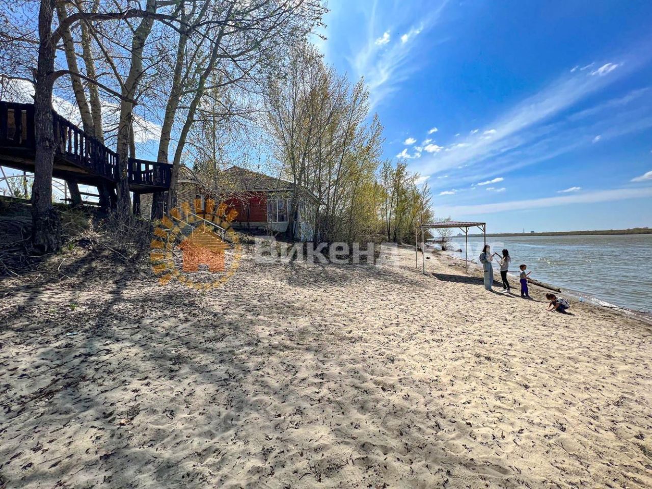 На фотографии видно песчаный пляж с рекой на переднем плане. Слева расположена деревянная терраса, возвышающаяся над уровнем земли, с перилами. На заднем плане виден дом с красной фасадной отделкой и крышей, наклоненной к спине. Вокруг растут деревья, создавая тень и уютную атмосферу. На пляже находятся люди, играющие и наслаждающиеся отдыхом. Цветовая палитра включает светлые песчаные тона и зелень деревьев, а также ярко-синий небосвод, что создает ощущение спокойствия и уединения.