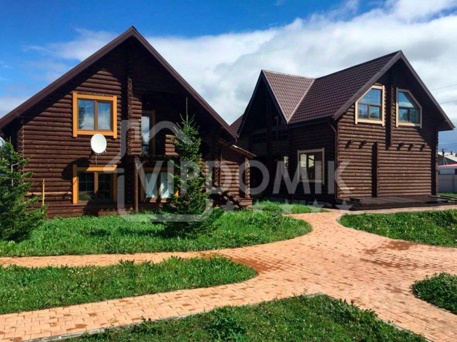 Архитектурный стиль изображает загородный дом в традиционном русском стиле с использованием деревянных бревен. Структурные особенности включают двускатную крышу и большие окна, обеспечивающие естественное освещение. На переднем плане виден ухоженный участок с тротуарной плиткой, ведущей к входу. Основные характеристики фасада – темное дерево с контрастными светлыми рамами окон. Цветовая палитра состоит из коричневых и светлых оттенков, создающих теплую атмосферу. Завершает общий вид аккуратный ландшафт с зелеными насаждениями, подчеркивающий уют и гармонию с природой.