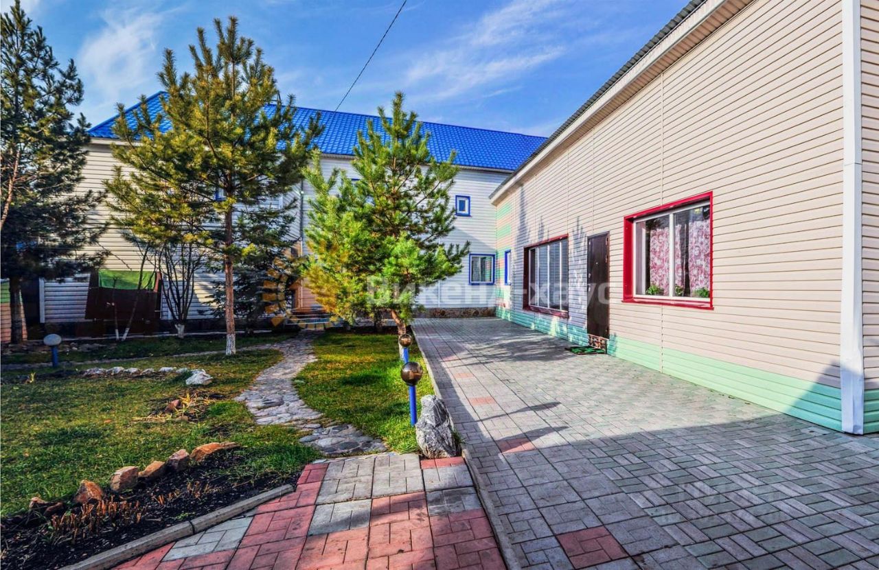 На фотографии представлено здание в современном архитектурном стиле с элементами минимализма. Фасад выполнен из светлого сайдинга с яркими акцентами на окнах и двери. Структурные особенности включают большие окна, обеспечивающие естественное освещение, и аккуратную планировку, которая создает ощущение открытости. Видно, что пространство вокруг вымощено плиткой, а также присутствует зеленая зона с небольшими деревьями и кустарниками. Основные цвета – светлые тона с яркими деталями, что придает помещению свежий и современный вид.