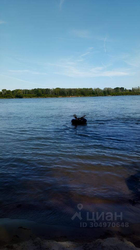 На фотографии видно спокойное водное пространство с мягкими волнами. На переднем плане находится резиновая лодка черного цвета, в которой сидит человек. Вдалеке виднеется зеленая растительность, образующая береговую линию. Вода имеет прозрачный синий оттенок, а небо ясное с легкими облаками. Цвета в изображении гармонируют, создавая атмосферу спокойствия и расслабления. Эта сцена может быть идеальной для отдыха на природе или активного времяпрепровождения на воде.