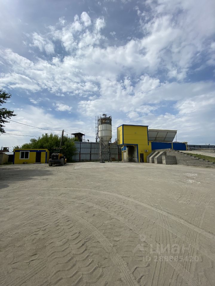 На фотографии представлено промышленное здание современного архитектурного стиля с ярко-желтой отделкой. Структурные элементы включают в себя большой резервуар для хранения и металлическую конструкцию с лестницей. Видна асфальтированная площадка, которая используется для маневрирования техники. Основные характеристики пространства включают просторный двор, окруженный высоким забором. Материалы, использованные в отделке, - бетон и металл, а цветовая гамма включает желтый и серый, что придает зданию современный и функциональный вид.