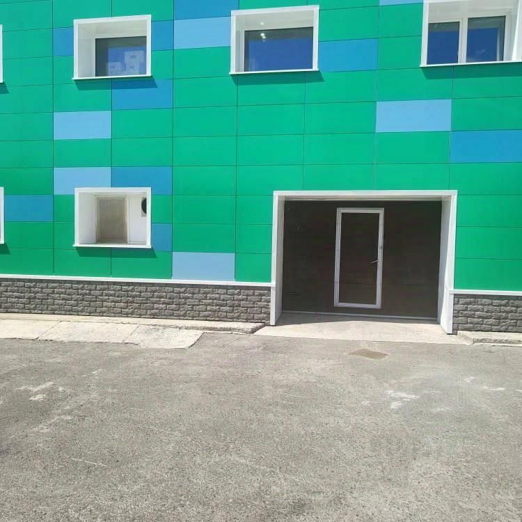 Фасад здания выполнен в современном стиле с яркой цветовой гаммой, где преобладают зеленые и голубые тона. Стена украшена плиткой, создающей эффект мозаики. На уровне первого этажа расположены окна с белыми рамами, а также входная дверь, ведущая в помещение. Дверь имеет стеклянные вставки и современный дизайн. Нижняя часть здания отделана серой каменной плиткой, что придает дополнительную текстуру и контраст. Общая композиция фасада создает впечатление свежести и современности.