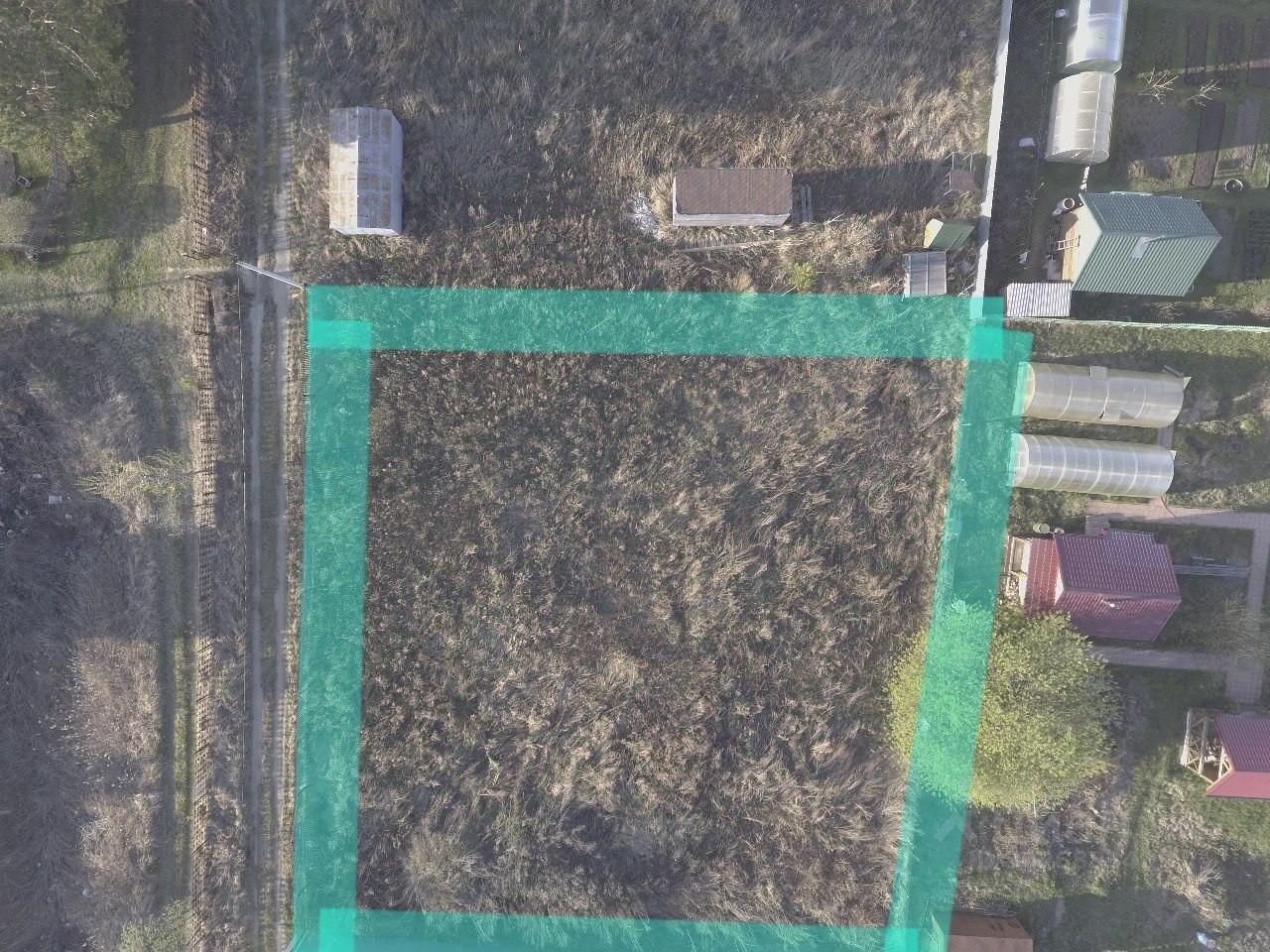 На фотографии представлено земельное участок, окруженное зелеными границами. Видно, что территория частично заросла травой и кустарниками. В верхней части изображения расположены несколько небольших строений, включая теплицы и дачные дома с красными и зелеными крышами. Дорога проходит вдоль участка, обеспечивая доступ. Структурные элементы включают в себя различные постройки, которые могут использоваться для хранения или отдыха. Общий вид участка создает ощущение уединения и возможности для садоводства или строительства.