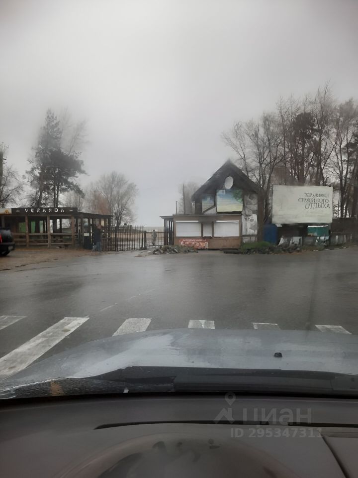 На фотографии виден деревянный дом с мансардной крышей и большими окнами, оформленный в загородном стиле. Стены здания частично окрашены, а также имеется вывеска с названием заведения. Вокруг расположены элементы благоустройства, такие как забор и дорожные знаки. Напротив дома находится открытая веранда с деревянной конструкцией, создающая уютную атмосферу. Основные цвета — коричневый и зеленый, что подчеркивает природный контекст. Пространство выглядит неухоженно, с некоторыми мусорными элементами на земле, что создает ощущение заброшенности.