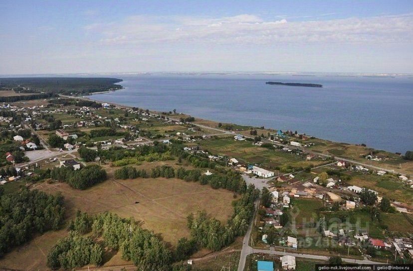 На изображении видно обширное пространство с зелеными насаждениями и участками земли, окруженными жилыми домами. Архитектурный стиль разнообразен, от одноэтажных до двухэтажных домов. Присутствуют асфальтированные дороги, которые соединяют разные участки. Цветовая палитра включает зеленые, коричневые и серые оттенки, отражающие природный ландшафт. Структурные элементы, такие как заборы и дорожные покрытия, придают месту ухоженный вид. Вдали виднеется водоем, что добавляет живописности этому району, создавая атмосферу спокойствия и уединения.