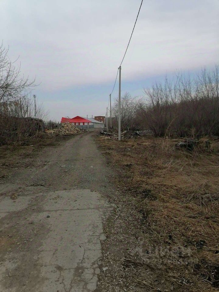 На фотографии виден грунтовый путь, ведущий к зданию с красной крышей. Дорога окружена участками с зарослями и строительными отходами. Слева расположены обломки и древесина, что придает местности заброшенный вид. Столбы электропередач тянутся вдоль дороги, создавая ощущение удаленности от городской инфраструктуры. Вдали виднеется еще одно строение, однако его детали неразличимы. Общая атмосфера изображения передает чувство уединения и частичной заброшенности пространства.