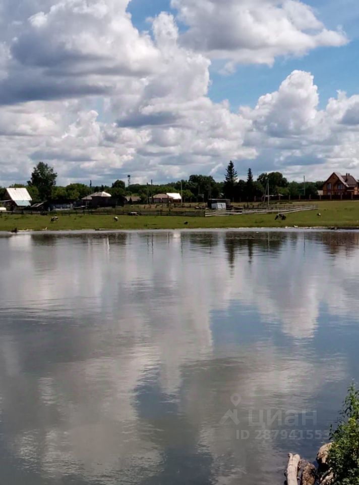 На фотографии виден водоем с спокойной водой, отражающей облака на небе. Береговая линия обрамлена зелеными насаждениями и кустарниками. На заднем плане расположены дома с различной архитектурой, среди которых выделяется деревянный дом с мансардой. Вокруг видны участки земли, вероятно, используемые для сельского хозяйства. Пейзаж создает атмосферу спокойствия и уединения, характерную для загородной жизни. Цвета в изображении преимущественно естественные: зеленый, коричневый и голубой, что подчеркивает гармонию с природой.