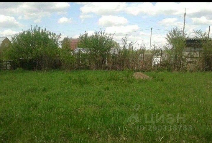 На фотографии представлено открытое пространство с зелёным травяным покрытием, окружённое кустарниками и небольшими деревьями. Видны участки с высокой травой, создающие естественный ландшафт. На заднем плане расположены строения, частично скрытые растительностью, что придаёт ощущение уединения. Пространство выглядит ухоженным, без крупных объектов, что позволяет сосредоточиться на природных элементах. Основные цвета – зелёный и коричневый, с разнообразием оттенков, в зависимости от освещения. Это место идеально подходит для создания сада или зоны отдыха.