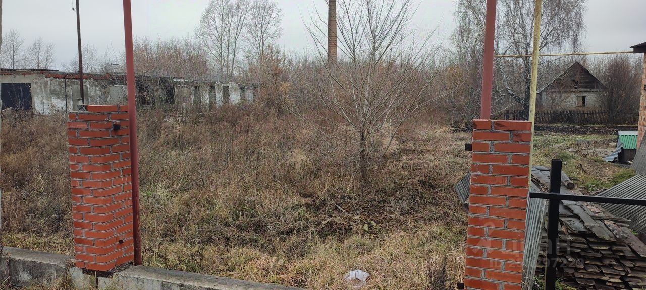 На фотографии виден участок земли с заброшенными строениями на заднем плане. Основные архитектурные элементы включают кирпичные столбы, которые создают ограничение пространства. Структурные особенности представляют собой старые стены и фрагменты зданий, частично разрушенные и покрытые плесенью. Основная зона изображения — это открытое пространство с высокой травой и кустарниками, что создает ощущение запущенности. Цвета доминируют серые и коричневые оттенки, а кирпичи имеют красный цвет. Общая отделка выглядит неухоженной, что подчеркивает заброшенность данного участка.