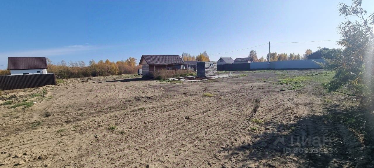 На фотографии виден участок земли с несколькими постройками. Архитектурный стиль домов простой, с двускатными крышами, отделанными черепицей. Участок имеет открытое пространство с ровной поверхностью, на которой видны следы от техники. Основные характеристики включают в себя несколько зданий, расположенных на расстоянии друг от друга, а также небольшую постройку, вероятно, для хозяйственных нужд. Цвета зданий нейтральные, с преобладанием белого и светло-коричневого оттенков. Огороженные участки создают ощущение приватности.