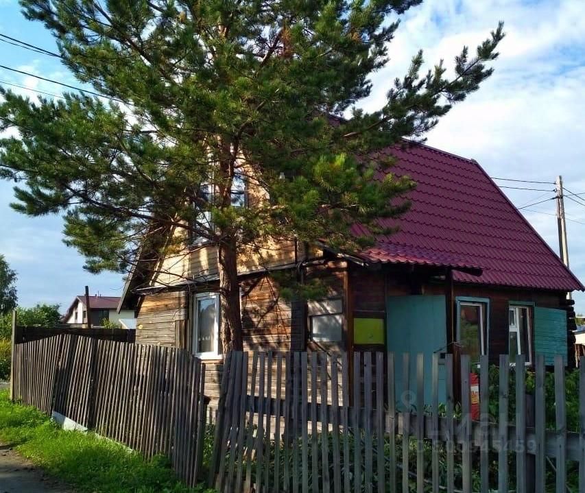 Дом в стиле дачи с деревянным каркасом и крышей из красной черепицы. Стены выполнены из бревен, что придаёт уютный вид. Окна имеют простые рамки, а одна из стен окрашена в светло-зелёный цвет. Вокруг дома установлен деревянный забор с горизонтальными планками, который обрамляет участок. На переднем плане виден зелёный газон и несколько высоких деревьев, создающих тень. Архитектурные элементы, такие как наклонная крыша и небольшие выступы, добавляют характер дому. Пространство выглядит гармонично и привлекательно для загородной жизни.