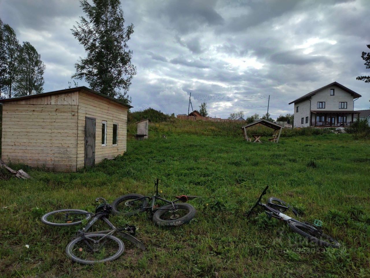 На фотографии представлено небольшое деревянное строение с простым архитектурным стилем. Стены выполнены из неокрашенного дерева, а крыша имеет плоскую форму. Окна здания квадратной формы, что придаёт ему минималистичный вид. Вокруг видны несколько велосипедов, лежащих на траве, что создаёт ощущение небрежности. На заднем плане расположено более крупное здание с большими окнами и балконом. Цветовая гамма в основном нейтральная, с преобладанием зелёных и коричневых тонов. Пространство выглядит открытым и естественным, с акцентом на деревянные материалы.