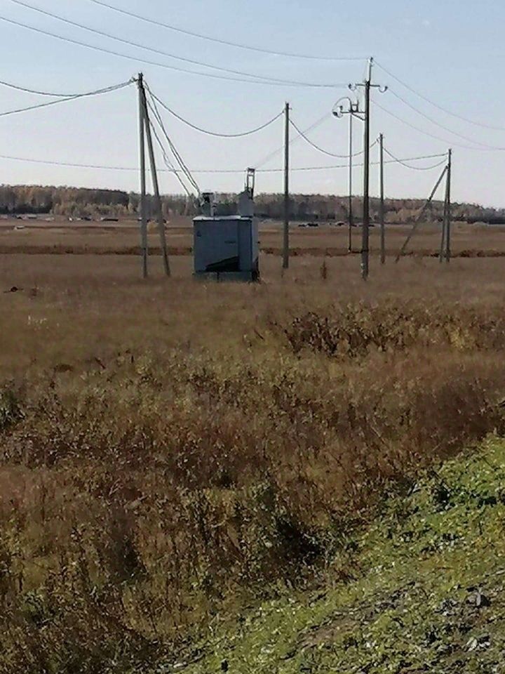 На изображении виден небольшой объект, возможно, трансформаторная подстанция, выполненный в простом современном стиле. Структура квадратной формы, окрашенная в светло-серый цвет с черными элементами. Вокруг видны электрические столбы с проводами, создающими сетевую инфраструктуру. Основные характеристики включают металлическую конструкцию и функциональный дизайн, обеспечивающий надежность. Пейзаж вокруг преимущественно открытый, с высохшей травой и редкими кустарниками, что подчеркивает удаленность объекта от городской застройки. Цвета нейтральные, с преобладанием серых и коричневых тонов.